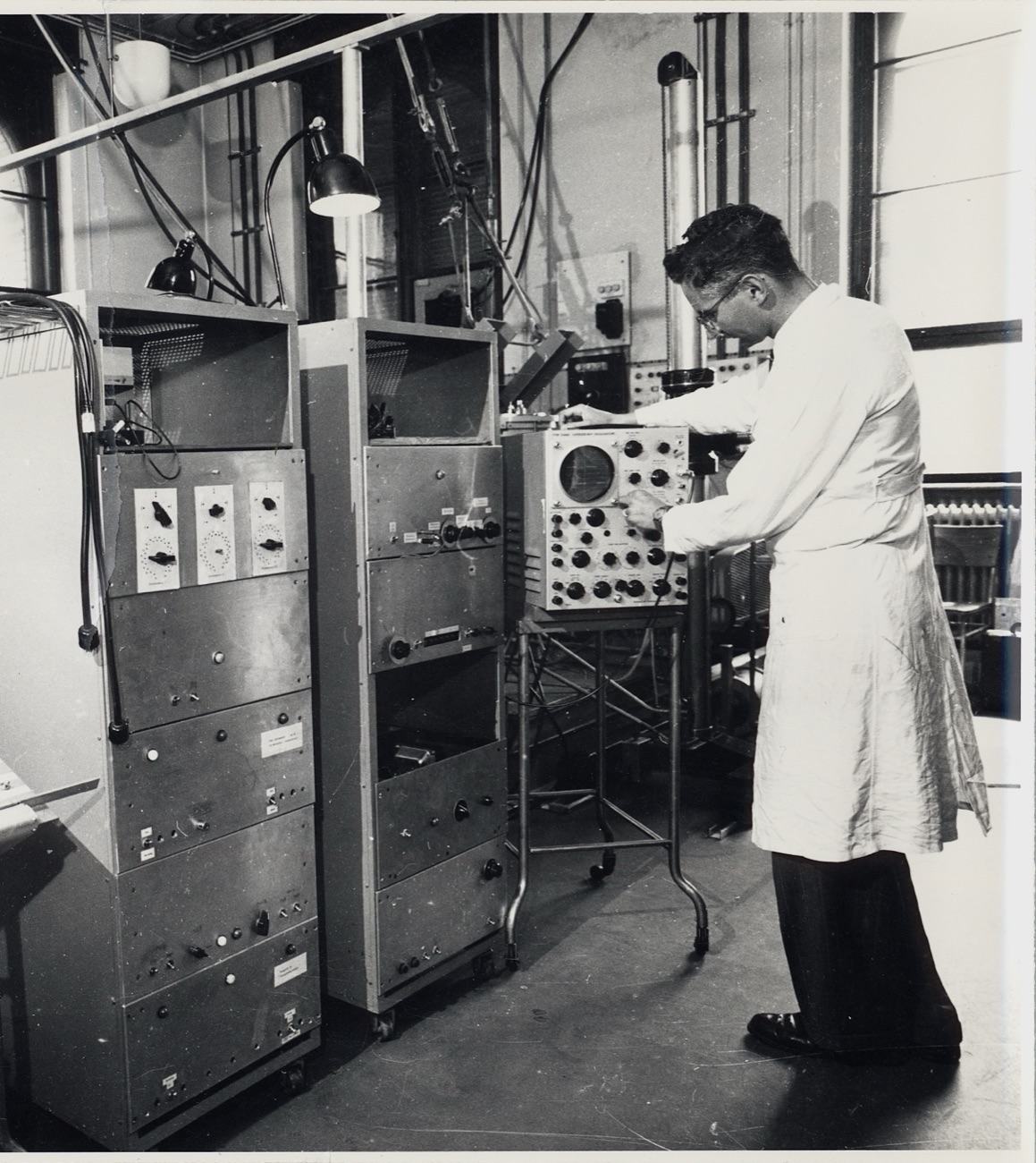 Photographie d'archive en noir et blanc : un technicien en blouse règle un oscilloscope au milieu des machines